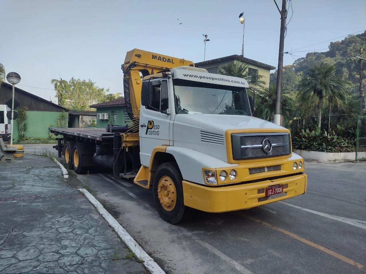 Caminhão Mercedes-Benz branco e amarelo do Peso Positivo com guindaste MADAL estacionado ao lado de vegetação no Zoológico de São Paulo