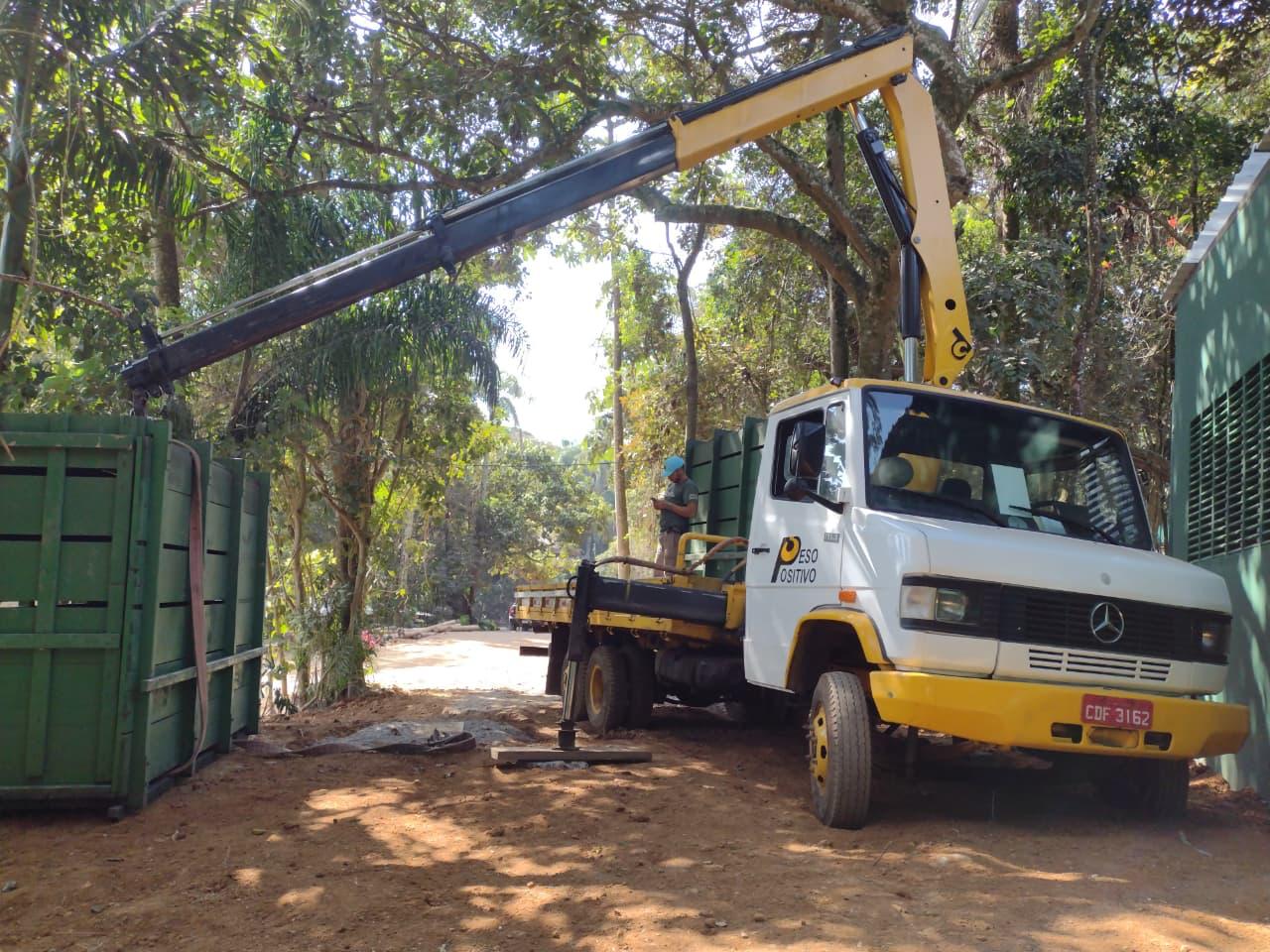 Caminhão Mercedes-Benz branco do Peso Positivo com munck amarelo estendido sobre caixa verde de girafa com cinta laranja em área de terra batida