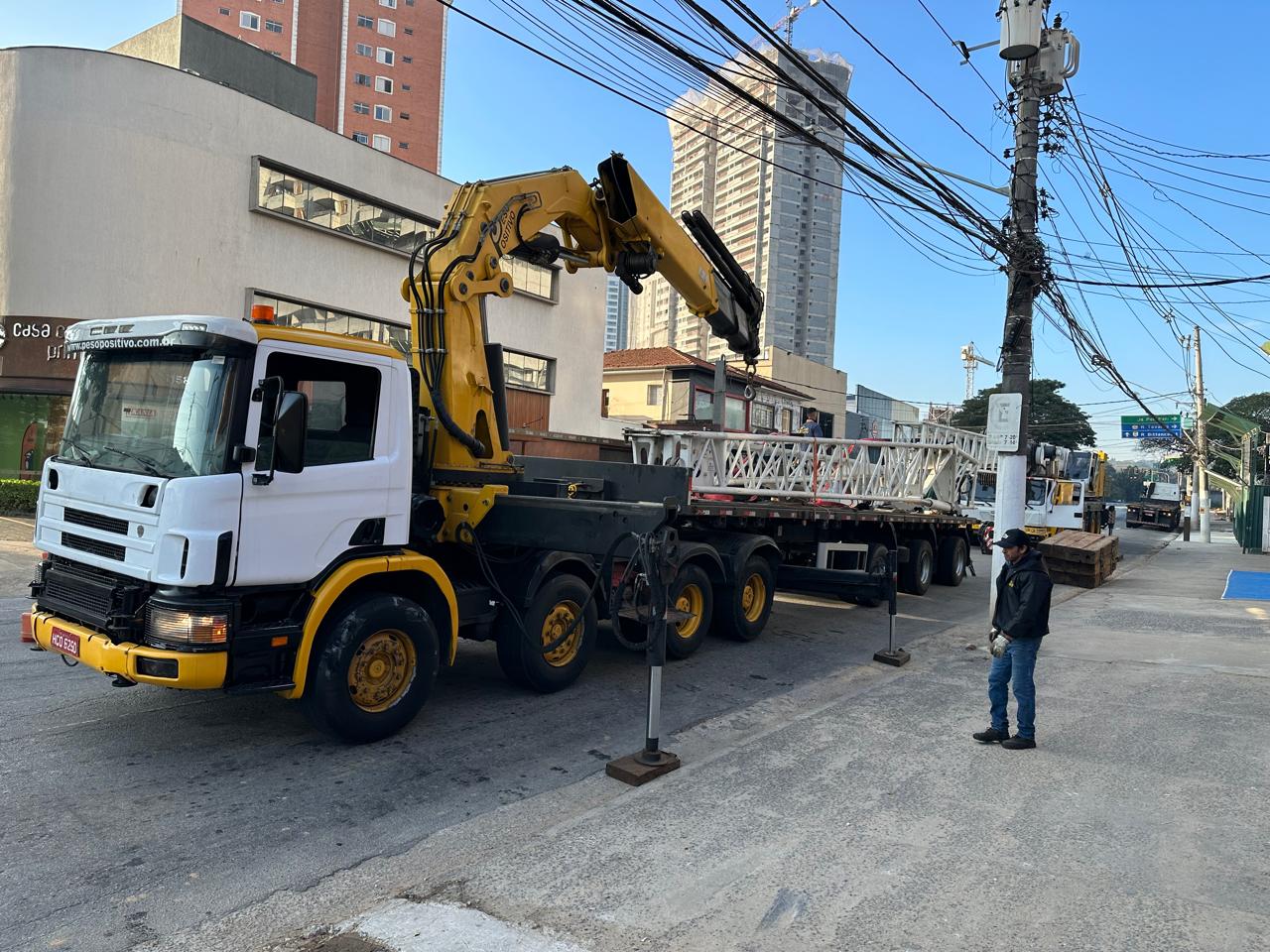 Caminhão Scania branco e amarelo do Peso Positivo com munck articulado e carreta carregada com seções treliçadas brancas da grua em rua urbana