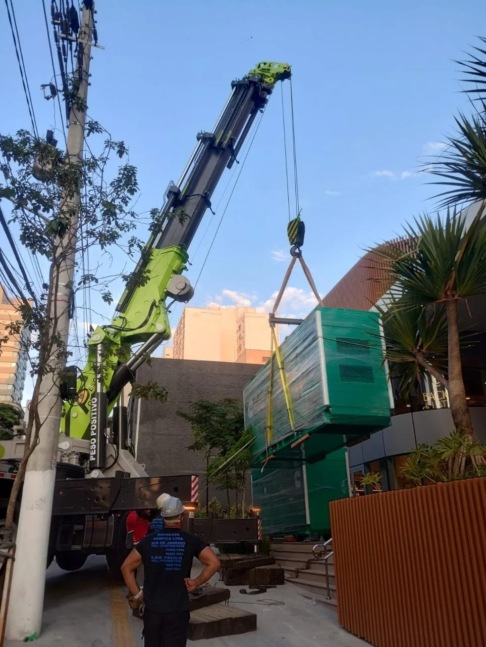 Gerador verde suspenso por cintas amarelas e balancim, início do içamento na rua