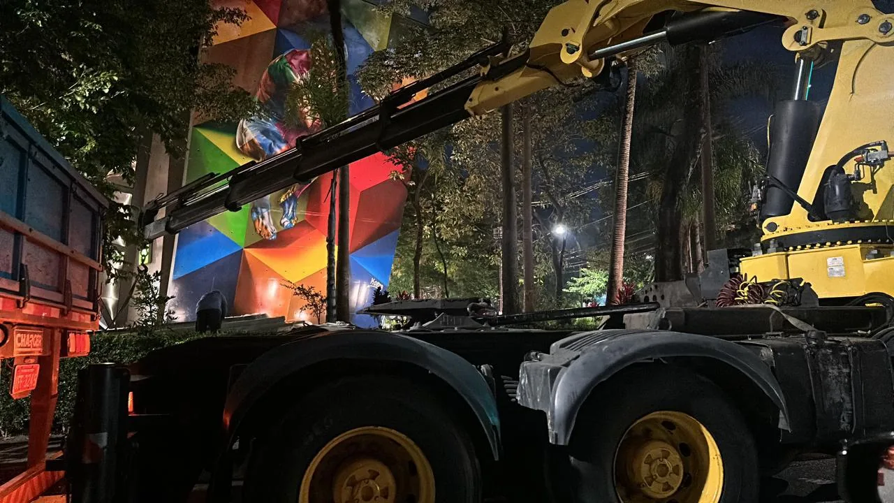 Braço do guindaste estendido em direção à fachada do prédio com mural geométrico, início do içamento das telhas