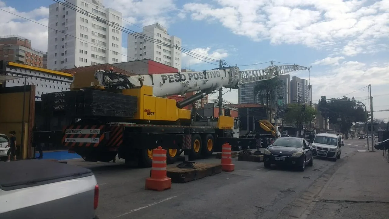 Guindaste Grove GMK 5275 amarelo e branco do Peso Positivo com patolas apoiadas em dormentes e jib treliçado, isolado por cones em via urbana