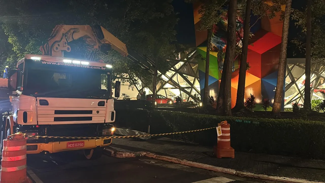 Caminhão com guindaste articulado estacionado na rua em frente à concessionária Ferrari nos Jardins, com cones e fita de isolamento durante a operação noturna