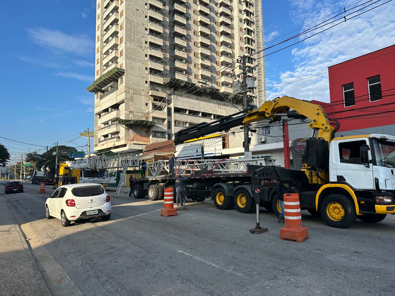 Camininhão Scania com munck articulado do Peso Positivo manuseando seções treliçadas brancas da grua ao lado do guindaste Grove em via urbana